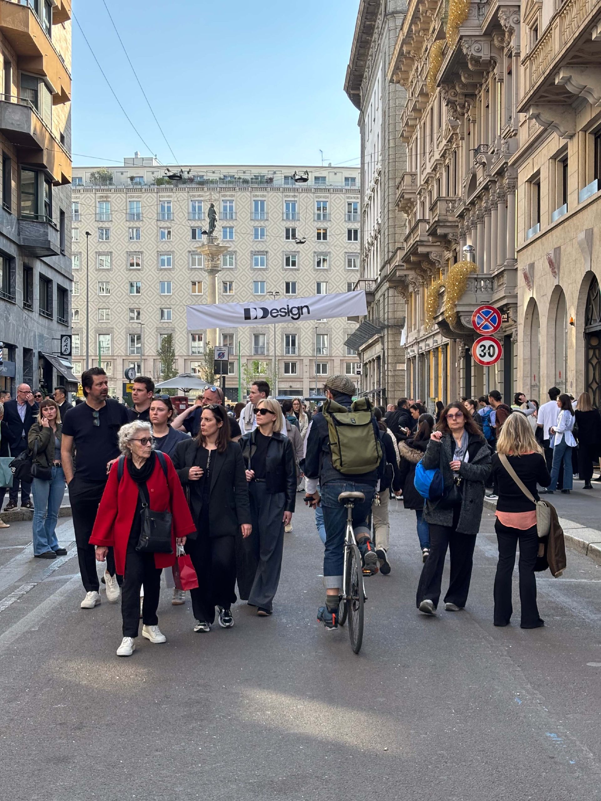 Folla a Milano durante la Design Week. Persone a piedi e in bicicletta in strada, banner con logo DDDesign. Architettura milanese sullo sfondo, evento di design, Fuorisalone, atmosfera vivace.
