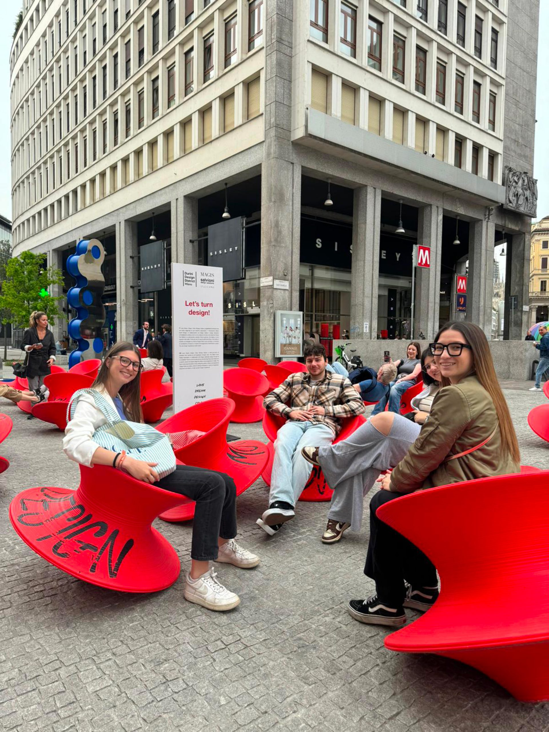 Installazione artistica di design a Milano: persone sedute su sedie rosse a forma di trottola in Piazza San Babila, vicino alla metropolitana. Design urbano e arte contemporanea a Milano.