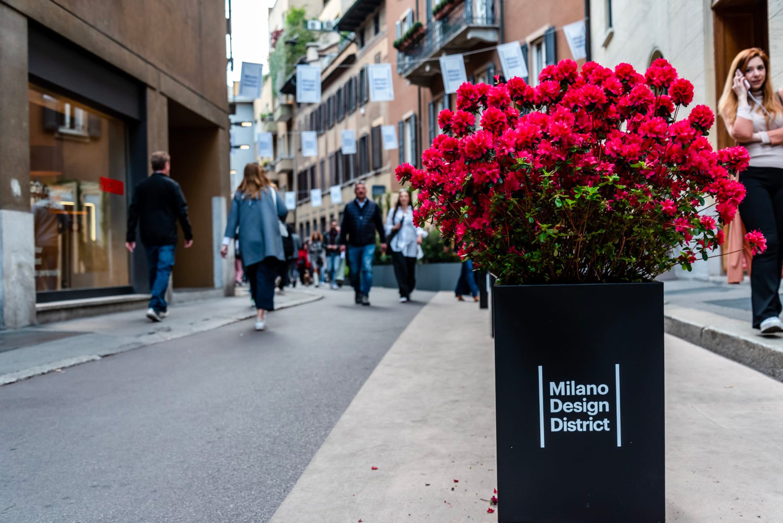 Milano Design District: Vaso di fiori rossi in strada durante la settimana del design. Architettura urbana, persone che camminano, bandiere decorative. Eventi e design a Milano.