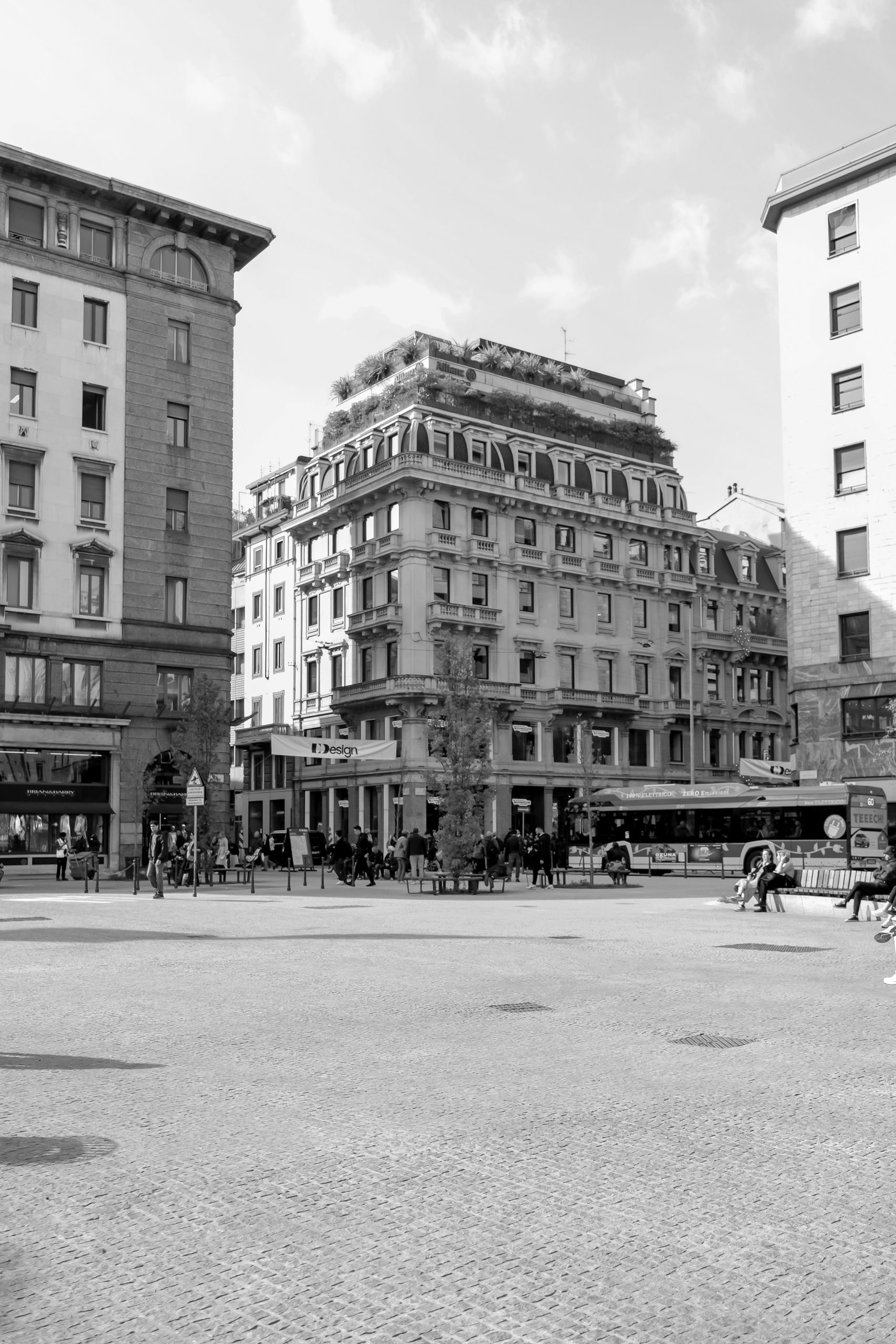 Piazza a Milano in bianco e nero. Architettura storica milanese, palazzi eleganti con dettagli classici. Persone sedute e in movimento, atmosfera urbana. Design e stile italiano.