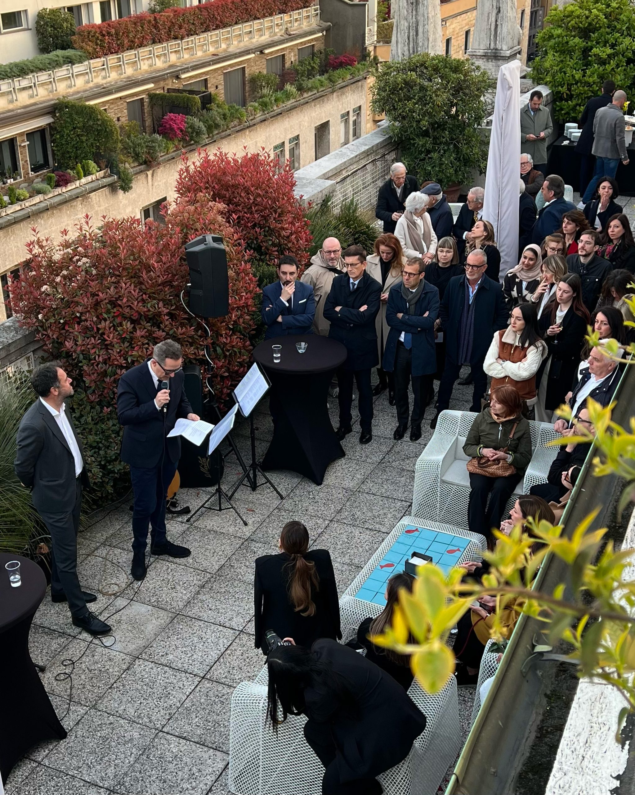 Evento rooftop a Milano: vista panoramica sulla città, networking e design. Persone che socializzano su terrazza con arredi moderni e vegetazione. Architettura milanese sullo sfondo.