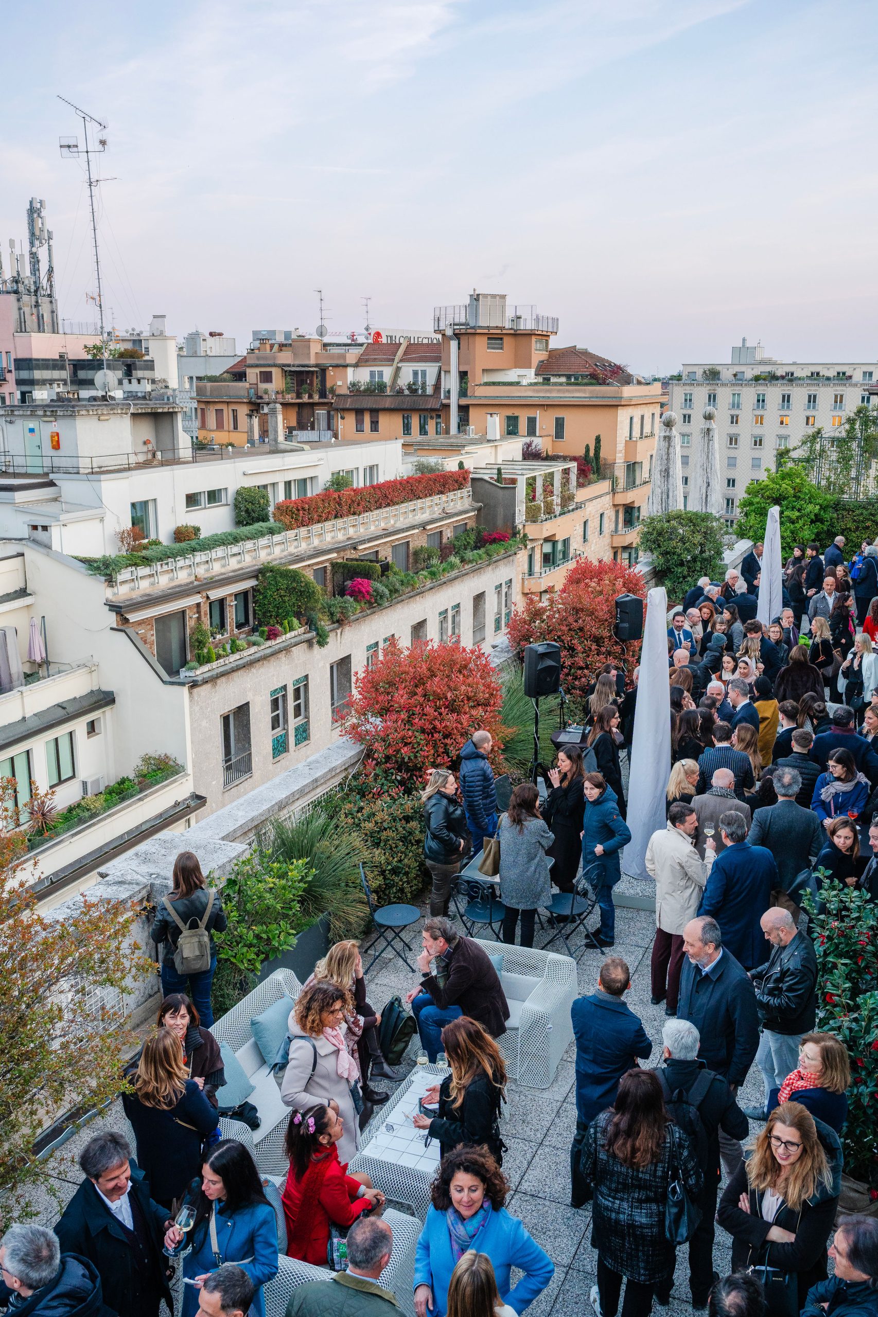 Evento rooftop a Milano: vista panoramica sulla città, networking e design. Persone che socializzano su terrazza con arredi moderni e vegetazione. Architettura milanese sullo sfondo.